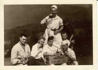 Black and white photograph of 7 people posing around a Artois crate.