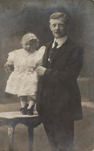 Photograph of a man posing with a girl in a dress standing on a table. - undated